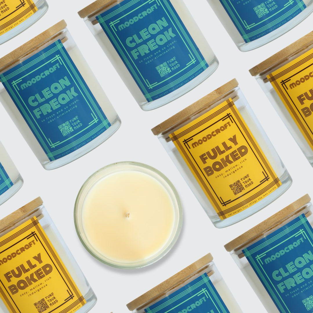 A styled flat-lay of multiple Moondrop candles on a white background, including several glass jars with blue and yellow labels and one open candle showing pale cream wax. The labels read 'moondrop' and the scent names 'CLEAN FREAK' and 'FULLY BOKED'.