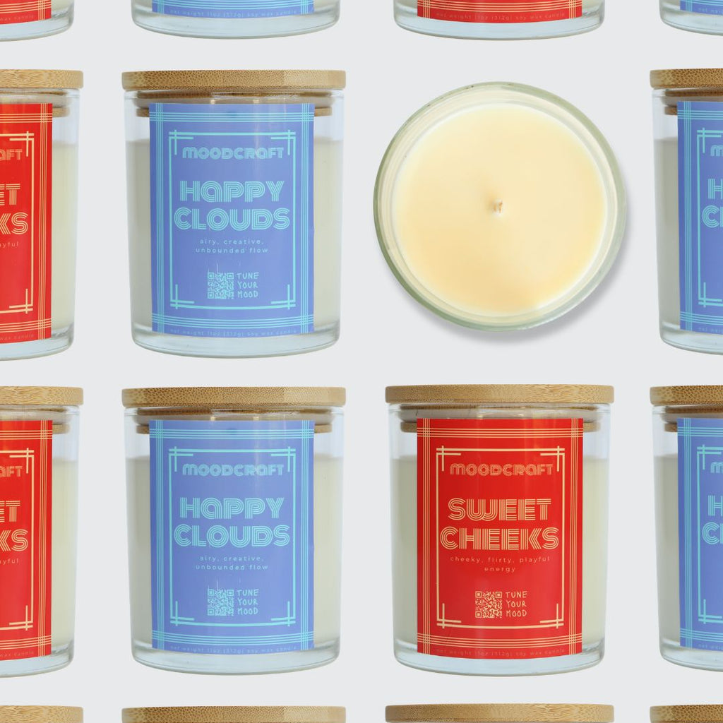 A repeating pattern of Moodcraft jar candles on a light gray background, with one candle shown open and centered to reveal the pale cream wax. The labels feature different scent names in red and blue, and the jars have light wood lids.