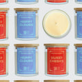 A repeating pattern of Moodcraft jar candles on a light gray background, with one candle shown open and centered to reveal the pale cream wax. The labels feature different scent names in red and blue, and the jars have light wood lids.