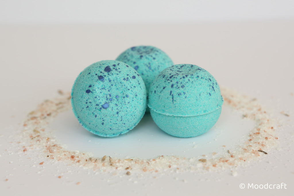 Three turquoise bath bombs with speckles on a white surface with sand-like material.